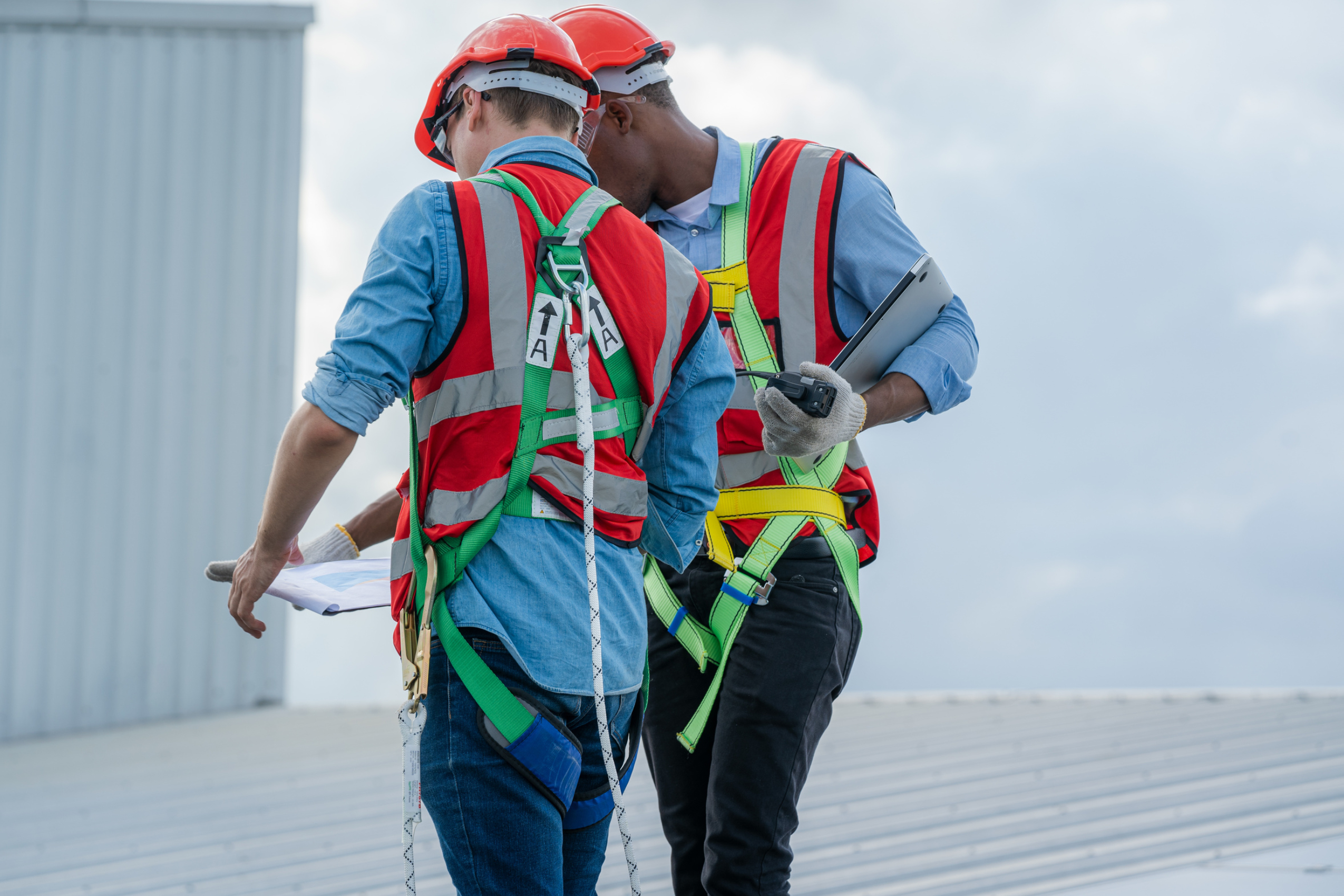 two male contractors with orange safety hats and safety harnesses on a roof having a discussion